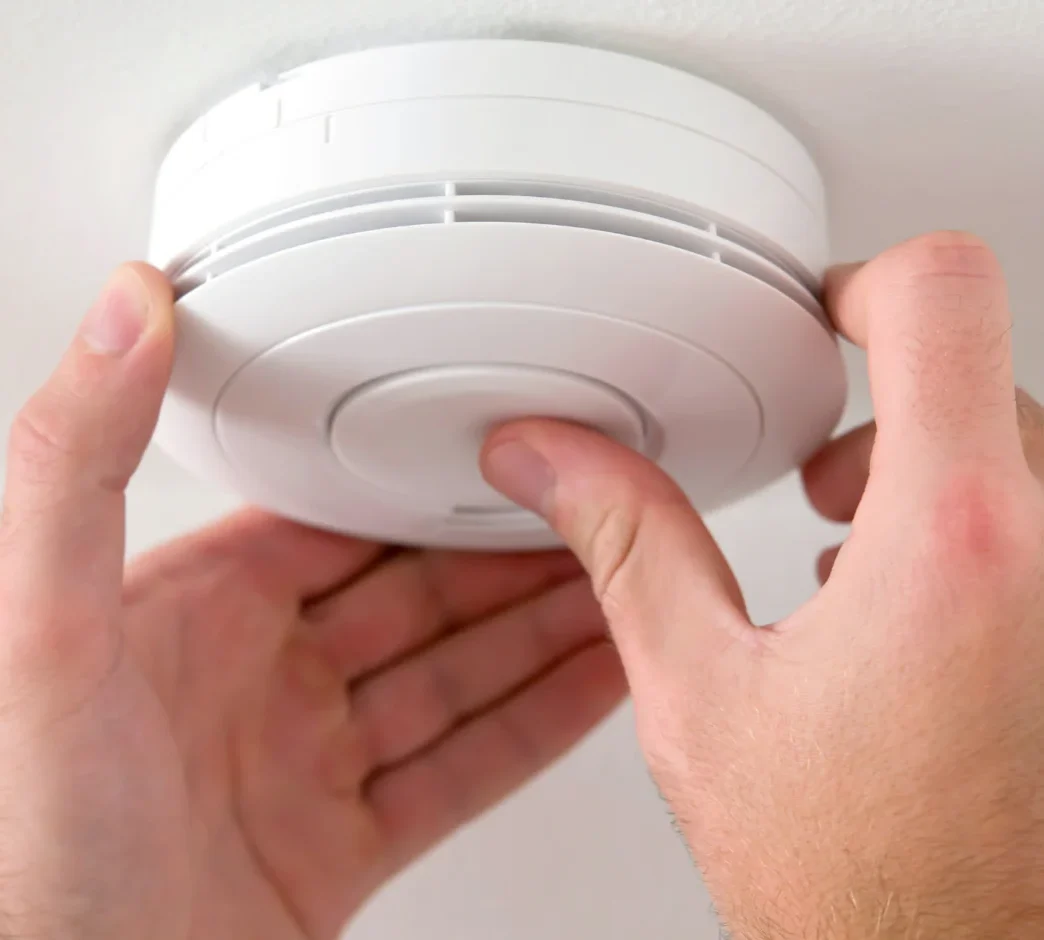 A person testing a white Carbon Monoxide detector mounted on a ceiling.