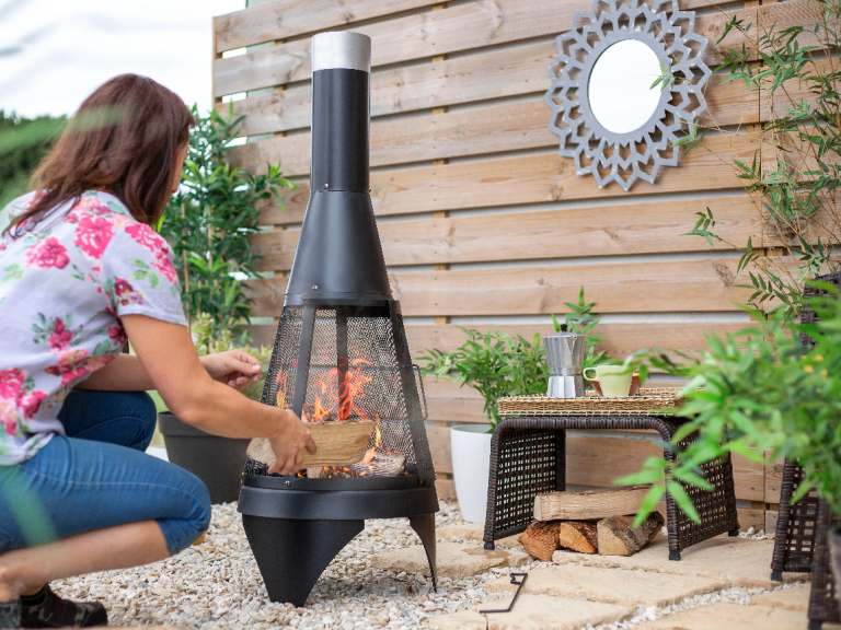 Woman tending a black outdoor fireplace in a garden setting.