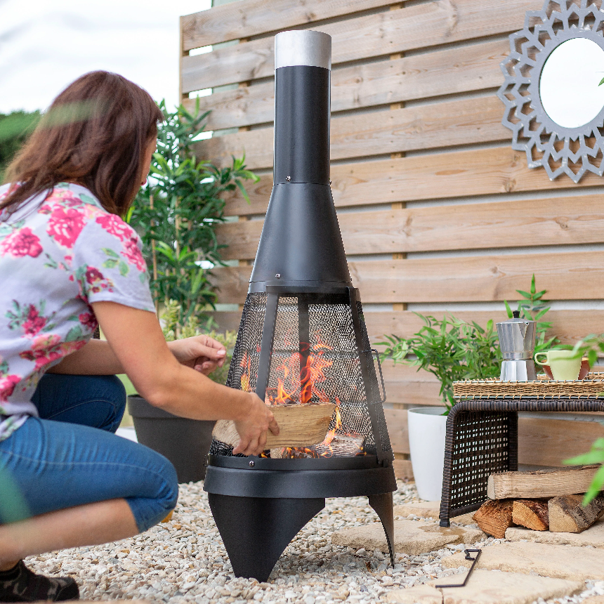 Woman adding wood to a black outdoor chiminea with plants and a wooden fence in the background.