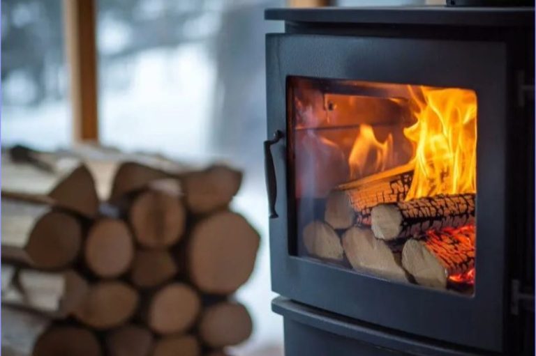 A wood-burning stove with flames and stacked firewood in a winter setting.