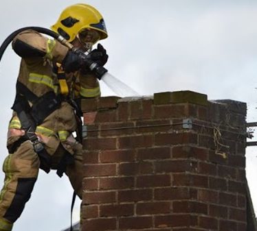 Firefighter extinguishing a chimney fire with a hose on a brick chimney.